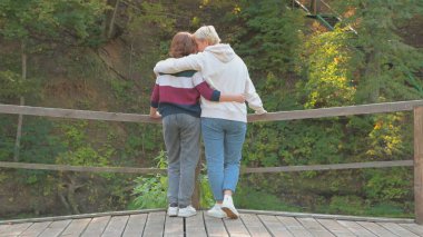 Anne ve kızı sırtı kameraya dönük bir şekilde doğada kucaklaşıyorlar..