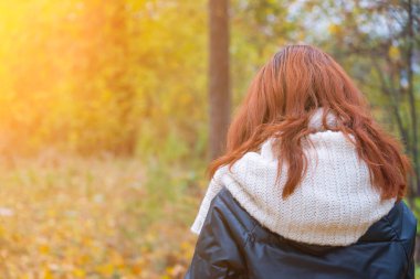 Siyah ceketli, kırmızı saçlı ve beyaz eşarplı bir kız sonbahar sarısı park manzarasında arkadan yürüyor..