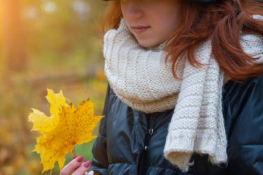 Ceketli, atkılı ve şapkalı, elinde akçaağaç yapraklarıyla gülümseyen kızıl saçlı bir kızın portresi. Sonbahar doğasının arka planına karşı, insani duygu kavramı.