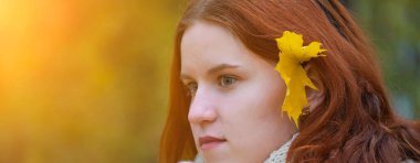 Kırmızı saçlı, ceketli ve atkılı, saçları açık ve kulağında akçaağaç olan gülümseyen bir kızın portresi. Sonbahar doğasının arka planına karşı, insani duygu kavramı.