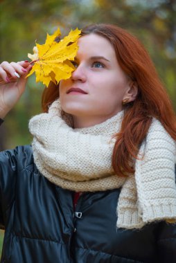 Elinde akçaağaç yaprakları olan, ceketli ve eşarplı kızıl saçlı gülümseyen bir kızın portresi. Sonbahar doğasının arka planına karşı, insani duygu kavramı.