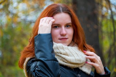 Ceketli, kızıl saçlı, gülümseyen bir kızın portresi elleriyle saçını düzleştiriyor. Sonbahar doğasının arka planına karşı, insan duyguları kavramı..