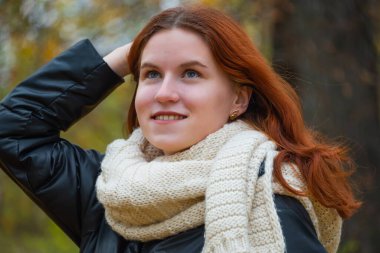 Ceketli, kızıl saçlı, gülümseyen bir kızın portresi elleriyle saçını düzleştiriyor. Sonbahar doğasının arka planına karşı, insan duyguları kavramı..