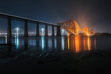 Gece vakti Forth Rail Köprüsü 'nün manzarası ve denizin üzerinde hareket eden bir trenin ışıltılı izi.