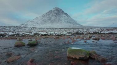 A calm river with stones on the foreground at the foot of a big mountain
