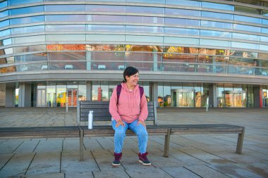 Kot pantolonlu, kapüşonlu, elinde demir bir şişe su olan genç bir esmer kız opera binasının önündeki ahşap bankta oturuyor. Kopenhag, Danimarka