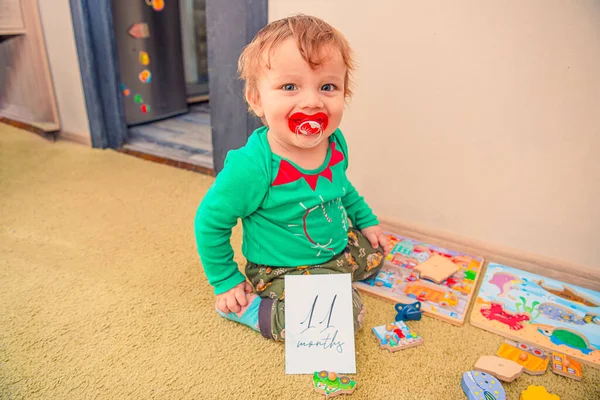 Ağzında bir emzik ve oyuncaklarıyla 11 aylık bir kilometre taşı kartı olan neşeli bir bebek.