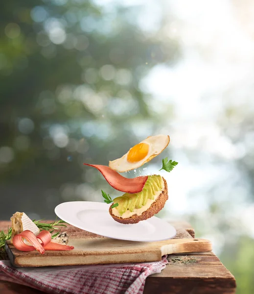 新鲜美味的自制三明治 配上鳄梨 烤鸡蛋和熏肉 在远离焦距的烤肉背景下 飘扬在木制桌子上 — 图库照片