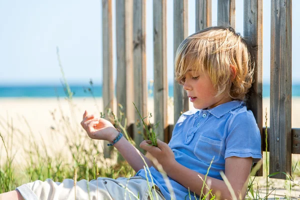 Chlapec sedí nex na dřevěný plot na pláži. — Stock fotografie