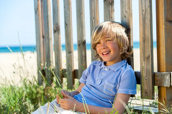 Lycklig pojke sitter bredvid stängslet på stranden. — Stockfoto