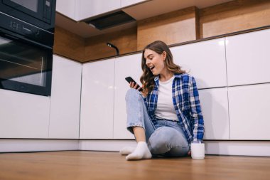 Mutfakta bir fincan çayla otururken cep telefonu kullanan mutlu genç bir kadının portresi..