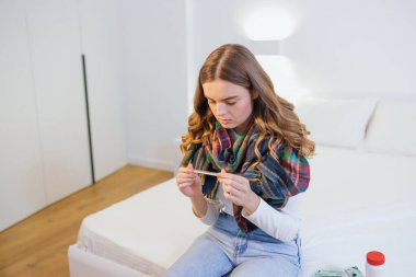 Yüksek ateşi olan bir yatakta oturan hasta bir kadın termometreye bakar. Soğuk ve migren..