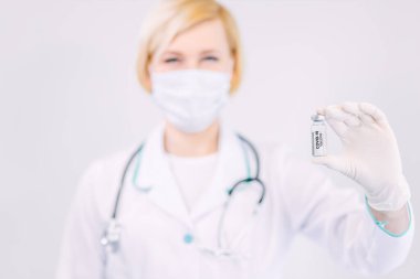Koruyucu maskeli ve beyaz önlüklü bir kadın doktor stüdyo arka planında tek başına dururken bir ampul COVID-19 aşısı taşıyor. Coronavirüs salgını konsepti