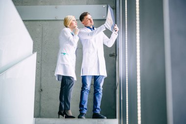 Modern bir tıp merkezinin koridorunda hastanın röntgenini çeken iki genç doktordan oluşan bir tıbbi ekibin yan görüntüsü. Hastalığın, viral hastalığın, akciğer sorunlarının gelecekteki tedavisi hakkında konuşuyorlar.