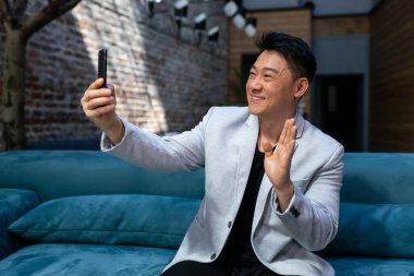 Neşeli Asyalı gülümseyen bir adam bir kafede oturmuş telefonda iş arkadaşlarıyla görüntülü konuşma yapıyor..