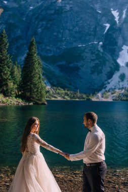 Güzel gelin ve damat dağlardaki gölün kenarında. Bir dağ manzarasının arka planında el ele tutuşup birbirinize nazikçe bakın.