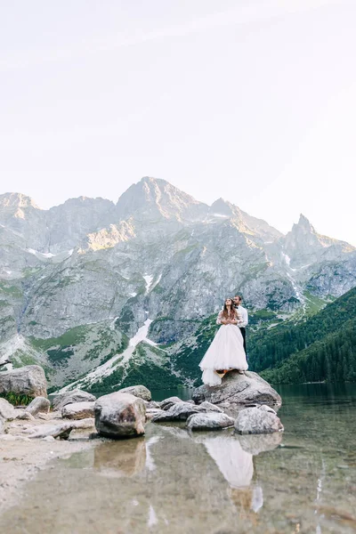 山の中の湖の近くの花嫁と新郎 山の風景を背景に新婚旅行を一緒に ロマンチックな結婚式のカップルの愛の石の海岸に立っている 絶景山景 — ストック写真