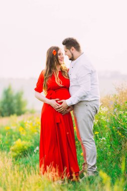 Genç hamile çift yaz günbatımında bir tarlada gülümseyip birbirlerine bakıyorlar..