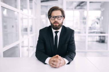 Ciddi sakallı ve gözlüklü bir adam modern beyaz ofis içinde masada oturuyor.