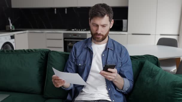 Man Works Smartphone Documents Couch Holding Paper His Hand Compares — Stockvideo