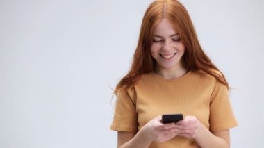 İzole edilmiş beyaz arka planda dururken akıllı telefon kullanan güzel bir genç kadın. Gülümsüyor, sosyal ağlarda cep telefonuyla iletişim kurarken eğleniyor.