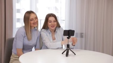 İki kız arkadaş el sallıyor, kalp yapıyor, abonelerini selamlıyor, webcam uygulamasında kameraya bakıyor rahat bir odada, oturma odasında, tıbbi karantinada, sosyal uzaklık izolasyonunda