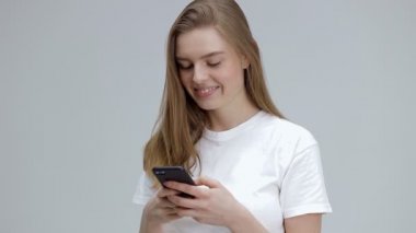 Tatmin olmuş bir kız cep telefonuna mesaj yazıyor, çevrimiçi iletişimden zevk alıyor, eleştiriler yazıyor, beyaz tişört giyiyor, gri bir arka planda izole edilmiş..