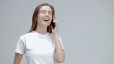 Heyecanlı genç bir kadın telefona cevap veriyor, gri stüdyo arka planında tek başına duruyor, mutlu genç bir kadın arkadaşıyla hoş sohbetler ediyor, akıllı telefon tutuyor, konuşuyor. Mutluluk duygusu