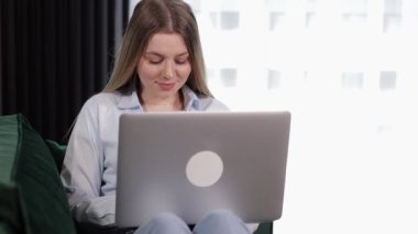 Kanepede oturan gülümseyen genç kadın dizüstü bilgisayar kullanıyor, ekrana bakıyor, mesaj yazıyor, mutlu kız internette, sosyal ağlarda video izliyor, evde çalışıyor ya da çalışıyor.