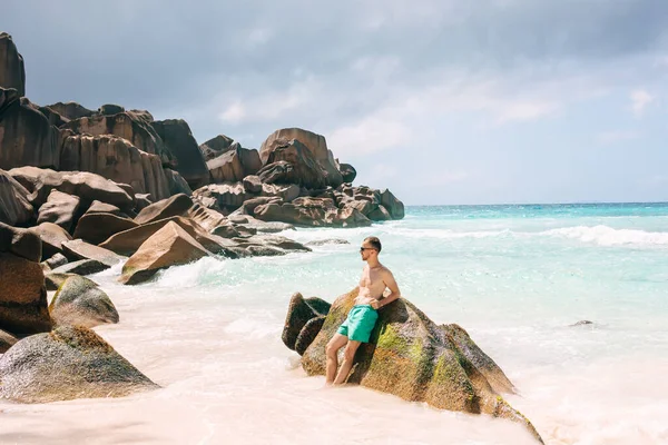 身穿泳裤的年轻人站在塞舌尔巴拉那热带岛屿的岩石海滩边 蓝蓝的天空和白云笼罩着 — 图库照片