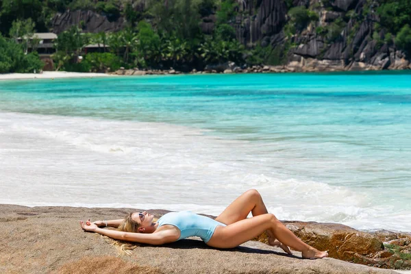暑假的概念 一个穿着蓝色比基尼泳衣 身材匀称 训练有素的性感美女躺在海滩上的石头上 在海滨摆姿势的时装模特 — 图库照片