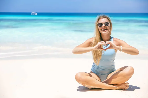 Cheerful woman in bikini sitting on the beach making heart shape with fingers. Woman by the sea in sunglasses.