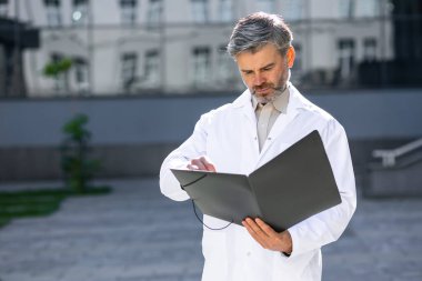 Elinde dosya olan bir erkek doktor bir hastanın raporuna ya da teşhisine bakar. Hastanenin arka planında durur.