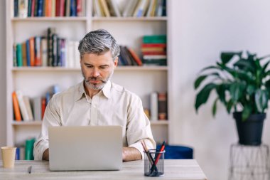 Ev ofisinin masasında oturan ve dizüstü bilgisayarda çalışan rahat giyimli yakışıklı bir adam.