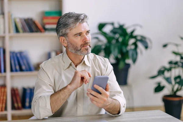 Hombre Serio Pensativo Mirando Distancia Sosteniendo Teléfono Inteligente Sentado Mesa —  Fotos de Stock