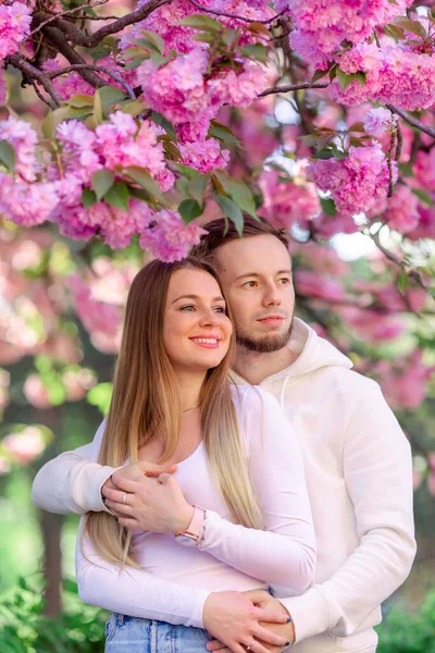 若いです幸せな笑顔ファッショナブルなカップルポーズ近くピンク春の花の木 — ストック写真