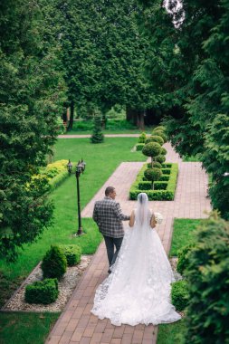Yeni evliler el ele tutuşup güzel bir parkta yürüyorlar. Arkadan bak. Uzun kuyruklu güzel gelinlik..