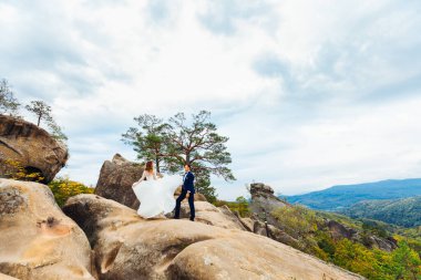 Gelinin elbisesi uçuyor. Dağların arka planında kayalıklarda yeni evliler
