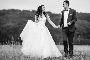 Gelin ve damat el ele tutuşup birbirlerine bakıyorlar. Siyah beyaz fotoğrafçılıkta, ormanın önündeki çimlerde yürüyorlar.