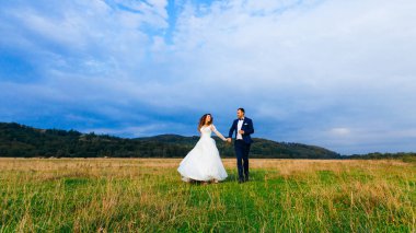 Şenlik kıyafetleri içinde yeni evliler el ele tutuşup ormanın yakınındaki çimlerde eğleniyorlar.