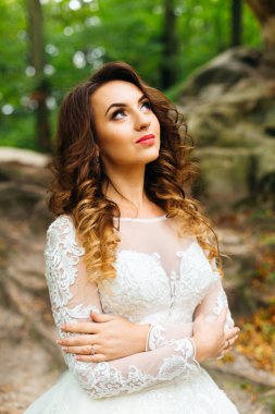 Bride in wedding dress hugging herself on the background of the forest and rocks