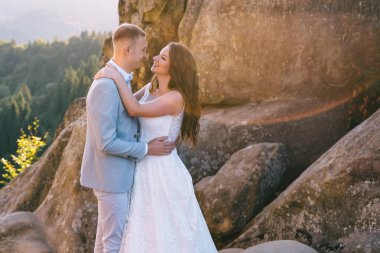 Sevgi dolu bir damat ve beyaz elbiseli güzel bir gelin kayaların arkasında bir kayanın üzerinde duruyorlar. Yeni evlilerin düğün fotoğrafı..