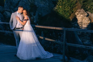 Yeni evlilerin dağlarda yürüyüşü. Uzun kıvırcık elbiseli yakışıklı gelin ve damat kayalardan bir arka planda sarılıyorlar..