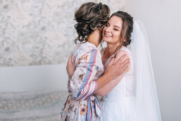 Bride closes her eyes hugging mother tender. Mom congratulates the bride with a marriage and hugs. Happy bride with mother on background wall in home. Bride morning preparation.