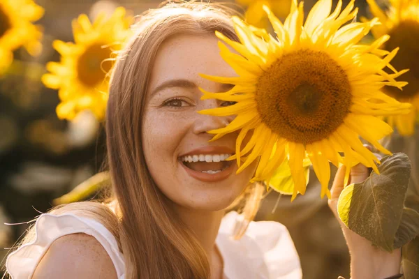 一个有着雀斑的快乐的女孩拿着一朵向日葵 在夕阳和田野的映衬下掩面 — 图库照片