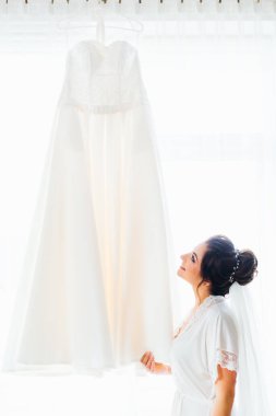 Güzel bir saç stili olan gelin, pencerenin arkasındaki gelinliğe bakar.