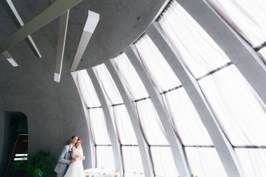 Damat gelini arkadan kucaklıyor büyük panoramik pencerelerin yanında. Restoran salonu