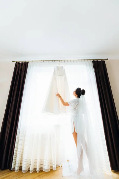 beautiful bride with bridal veil touches the wedding dress on the background of a window with a tulle and a curtains. beautiful hairdo