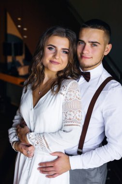 The guy hugs the girl from the back side and they look at the camera. white festive clothing