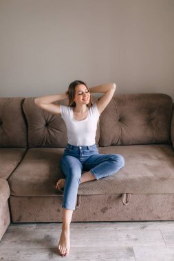 In cozy living room happy woman put hands behind head sitting leaned on couch